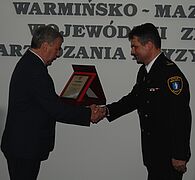 Na zdjęciu dwie osoby. Z lewej wojewoda warmińsko-mazurski Marian Podziewski wręcza certyfikat komendantowi Straży Miejskiej, Jarosławowi Lipińskiemu. Panowie podają sobie ręce.