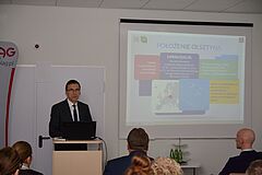 Po lewej przy mównicy prezydent Olsztyna Piotr Grzymowicz podczas prezentacji miasta na spotkaniu z przedsiębiorcami. Z prawej na ekranie wyświetlona prezentacja.