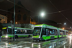 Na pierwszym planie stojący na światłach tramwaj. Obok, po lewej stronie zdjęcia autobus.