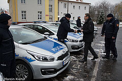 Prezydent Olsztyna Piotr Grzymowicz przekazuje nowe radiowozy. Auta ustawione są w rzędzie po lewej stronie. Policjant ściska dłoń prezydenta