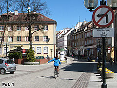 Rowerzystka podróżująca uliczkami Starego Miasta. Po obu stronach brukowanej ulicy zaparkowane auta.